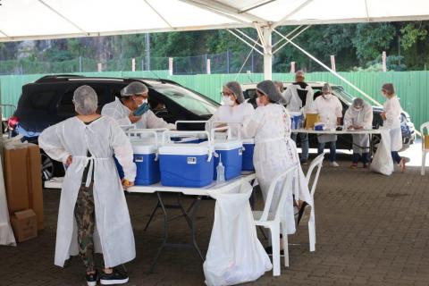 Drive thru movimentado por pessoas vestidas de branco. Sobre as mesas há coolers onde estão as vacinas. #paratodosverem