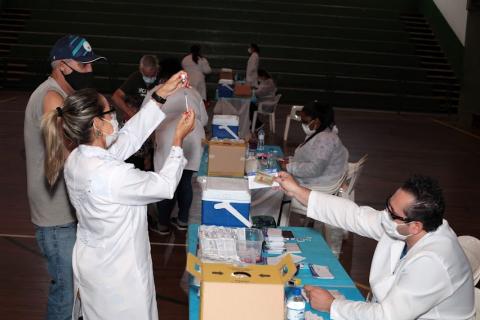 equipe de atendimento está em torno de uma mesa. Em primeiro plano uma  mulher segura uma seringa. #paratodosverem