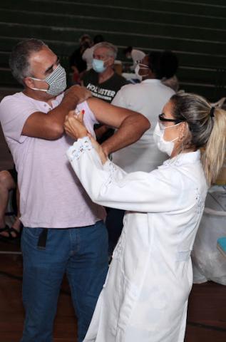 Homem está em pé, usando máscara e sendo vacinado por uma mulher. #paratodosverem