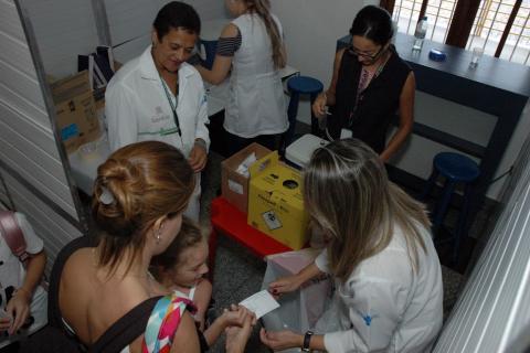 mulher e criança recebem carteirinha de vacinação. Foto é feita do alto. A mãe está de costas e a criança aparece abaixo. Há quatro pessoas envolvidas no atendimento. #paratodosverem