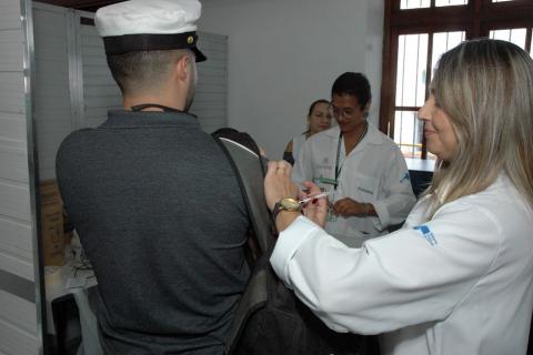 mulher aplica vacina em homem que usa chapéu de comandante de navio. #paratodosverem
