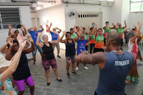 mulheres fazem movimentos de dança no salão orientadas por professor que está de costas para a foto. Todas elas estão com os braços para cima. #paratodosverem