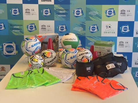 bolas para esporte, uniforme, cones e caneleiras sobre uma mesa. #paratodosverem