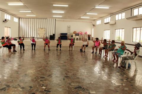 salão com várias mulheres sentadas em cadeiras e fazendo movimentos com os braços. #paratodosverem