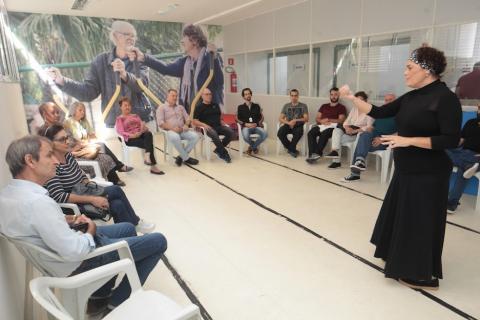 várias pessoas sentadas e uma em pé falando a todos. Ao fundo, parede é decorada com grande painel onde se vê dois idosos fazendo exercícios. #paratodosverem