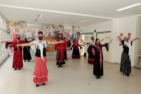 mulheres usando roupas típicas do flamenco, com saias rodadas, dançam em salão. Todas estão com os braços abertos. #paratodosverem