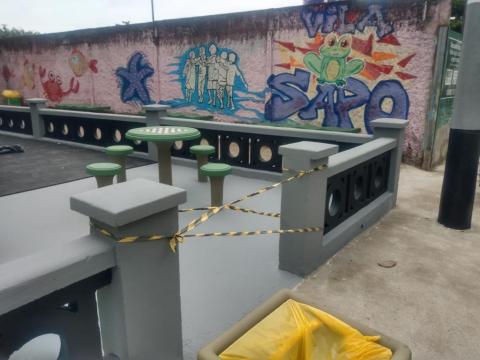 Espaço com muro colorido, mesas e entrada bloqueada por fita. #paratodosverem