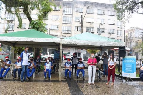 tendas montadas em praça com pessoas no interior. Duas mulheres estão à frente, uma delas fala ao microfone. #paratodosverem