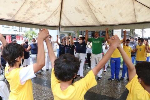 Pessoas estão sob tenda, de mãos dadas para o alto. #paratodosverem