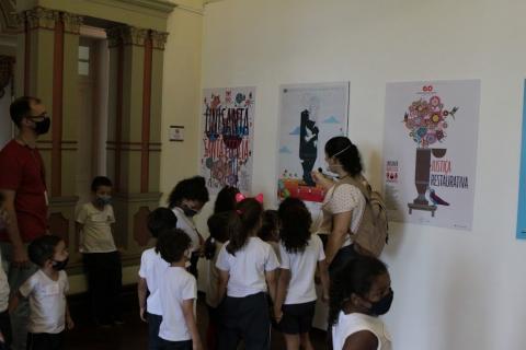 crianças olham para três obras afixadas em parede. Professora orienta o grupo. Também há um homem acompanhando. #paratodosverem