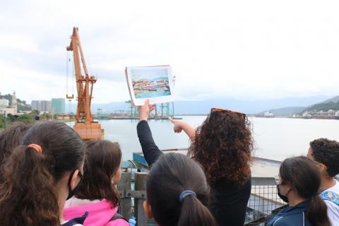 crianças estão de costas para a foto olhando foto que a professora segura. A imagem na mão dela é a mesma que eles veem ao vivo e ao fundo, isto é, uma vista geral do porto.  #paratodosverem