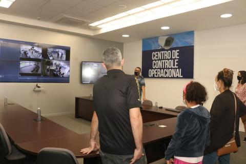 pessoas estão em pé em sala de reunião. Ao fundo, placa afixada tem os seguintes dizeres Centro de Controle Operacional. #paratodosverem