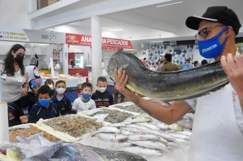 crianças estão diante de caixas cheias de peixes e olhando para homem que segura um peixe grande, em primeiro plano. #paratodosverem
