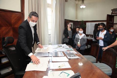 estudantes estão na sala do gabinete do prefeito. Ele está em pé, mexendo em papeis sobre a mesa. #paratodosverem