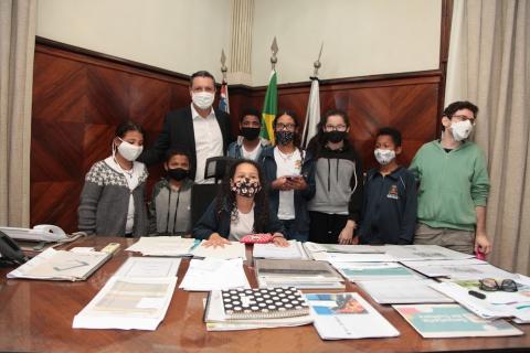 Estudantes estão no gabinete do prefeito. Uma delas está sentada à mesa do chefe do executivo. Todos posam para foto. #paratodosverem