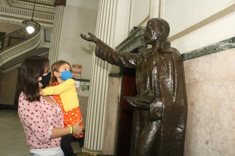 Mulher segura criança no colo. Ambas olham para a estátua do padre José de Anchieta. #paratodosverem