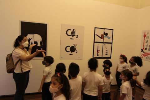 crianças observam três obras expostas. A professora faz gestos explicando para a turma. #paratodosverem