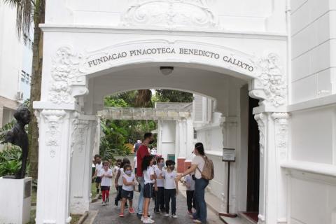 crianças e dois adultos se preparam para entrar na pinacoteca. No alto da entrada do casarão se lê Pinacoteca Benedicto Calixto. #paratodosverem