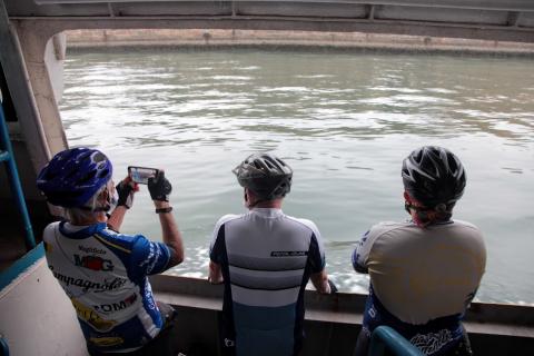 dentro da barca, três ciclistas olham o mar Um deles fotografa com o celular. #paratodosverem