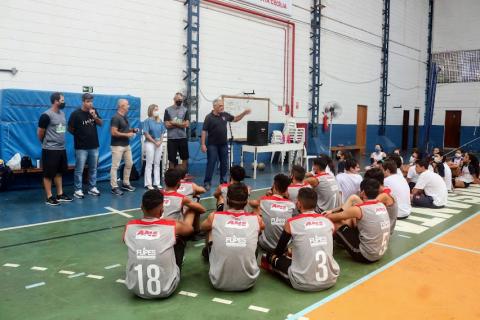 alunos e atletas estão sentados ouvindo um homem que está em pé falando. ele está ao lado de outros. Todos estão em quadra poliesportiva. #paratodosverem