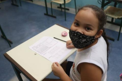 menina está sentada com papel à frente. Ela olha para a câmera. #paratodosverem