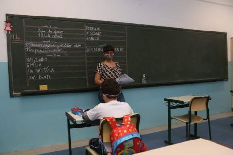 professora está em pé à frente de lousa. Um aluno está sentado na carteira escolar de costas para a foto. #paratodosverem