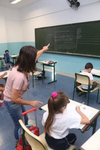 professora está em pé apontado para a lousa. Ela está ao lado de uma aluna sentada na carteira escolar. #paratodosverem