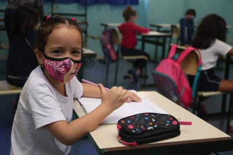 memina está sentada na carteira escola. Ela está de máscara e olha para a câmera. #partodosverem