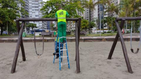 Homem realiza reparo em cima de balanço de playground na orla #paratodosverem