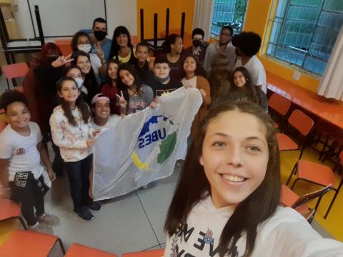 Estudante faz selfie com outros alunos com bandeira ao fundo #paratodosverem