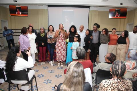 mulheres e homens reunidos em salão. Uma delas posa com troféu. #paratodosverem