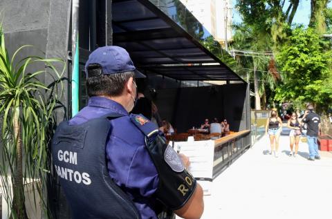 Guarda municipal está de costas, fazendo anotação em papel sobre prancheta. Ele está na rua, ao lado de um bar de portas abertas. #Paratodosverem