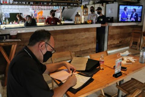 Fiscal está sentado e escreve emum papel sobre uma mesa. Ao fundo há homens trabalhando atrás de um balcão de bar. #Paratodosverem