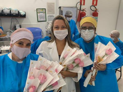 Mulheres usando avental, touca e máscaras carregam rosas nos braços. #Paratodosverem