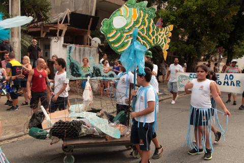 Carrinho com resíduos descartáveis, crianças fantasiadas, carregado peixes estilizados. #Paratodosverem