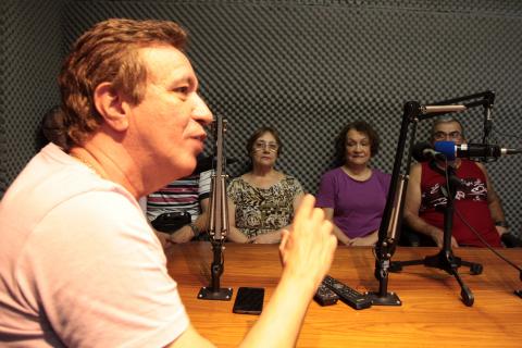 grupo está sentado na mesa da rádio com microfones no meio #pracegover 