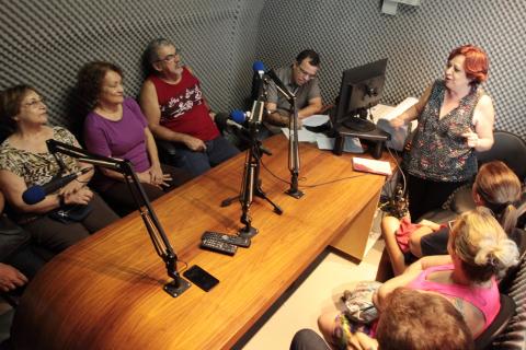 grupo está sentado na mesa da rádio com microfones no meio #pracegover 