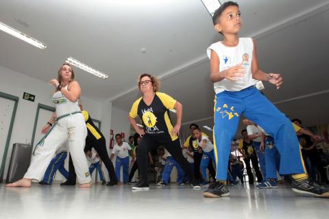 Menino, em primeiro plano, faz movimentos de capoeira. Atrás, duas mulheres e outras crianças acompanham. #Pracegover