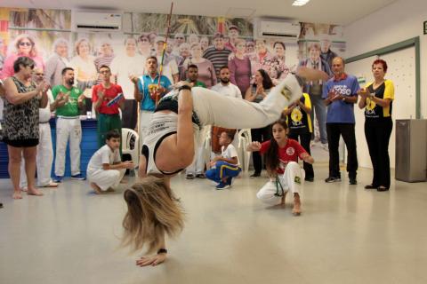 Jovem está apoiada no chão com somente uma das mãos. Seus cabelos cobrem seu rosto. Ao fundo, os integrantes da roda de capoeira tocam instrumentos e batem palmas. #Pracegover