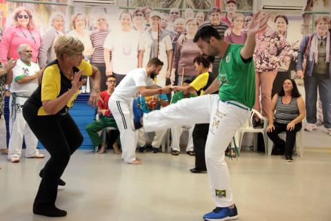 Idosa e jovem jogam capoeira em salão. Outras duplas também praticam. #Pracegover