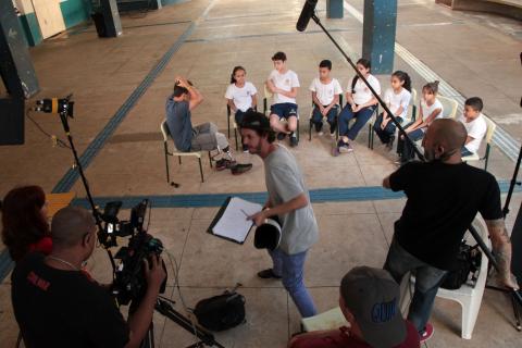 Conversa de Pauê com alunos é grava por equipe em área livre da escola. O entrevistado e as crianças estão sentados em semi-círculo. A equipe de gravação está em pé. Há cinco pessoas. Elas manuseiam câmeras, microfones e outros equipamentos. #Pracegover