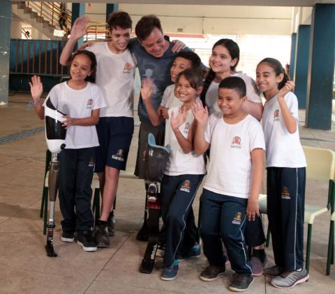 Pauê está em pé posando para foto com as crianças ao lado dele. As próteses estão à frente de duas crianças. #Pracegover