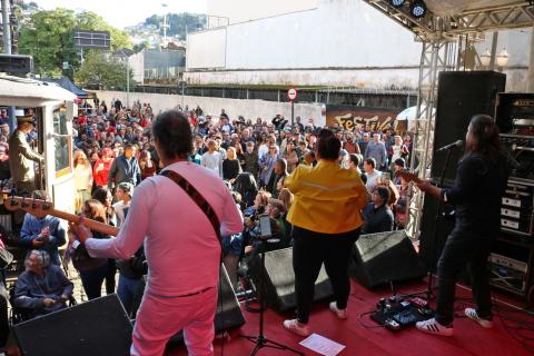 Três músicos estão se apresentando em palco. Ao fundo, o público acompanha. #Pracegover