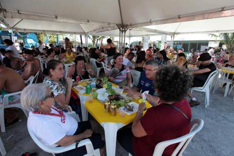 #pracegover Sob tenda, cerca de 50 pessoas sentadas junto a mesas comem e bebem
