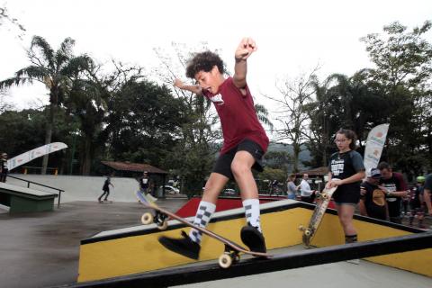 Menino faz manobra em rampa da pista. Atrás, menina com skate na mão se prepara manobra