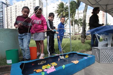 Crianças brincam de pescaria diante de pequeno tanque. Elas seguram pequenas varas e tentam pescar reproduções de peixes. #Pracegover