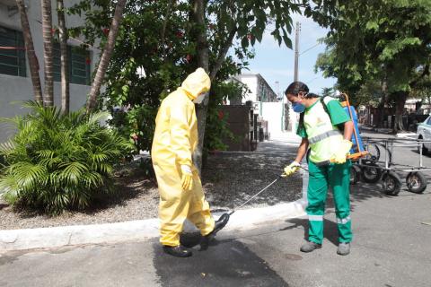 Mulher usando luva e máscara esguicha produto em homem que está usando macacão, bota, luvas, gorro e máscara