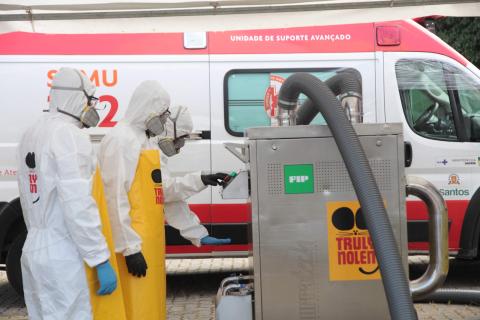 operários protegidos olham para máquina de limpeza com ambulância ao fundo #paratodosverem