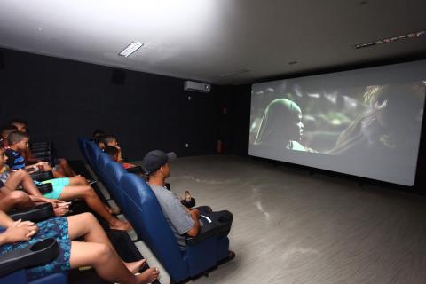 Sala de cinema lotada de jovens. Eles estão sentados olhando para telão à frente. #Pracegover