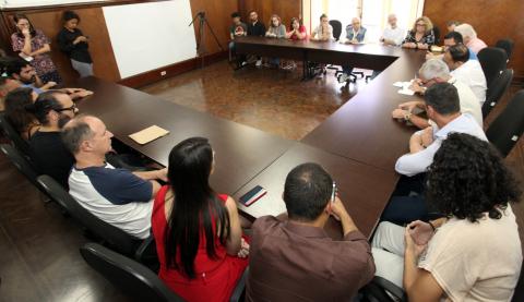 Pessoas sentadas em mesa durante lançamento do concurso #pracegover 